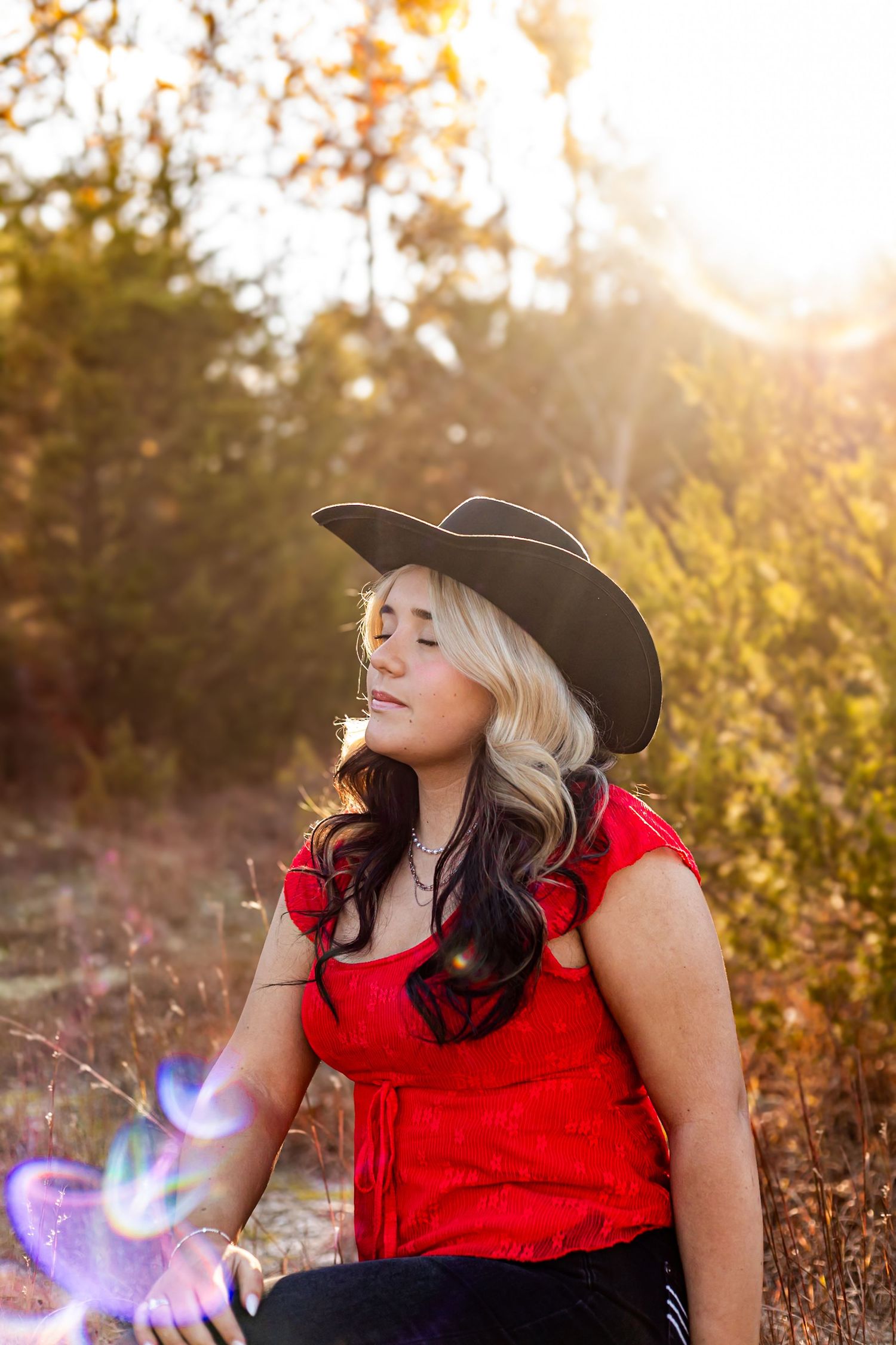 Blonde woman in black cowboy hat and red top sitting outdoors in golden sunlight among trees.