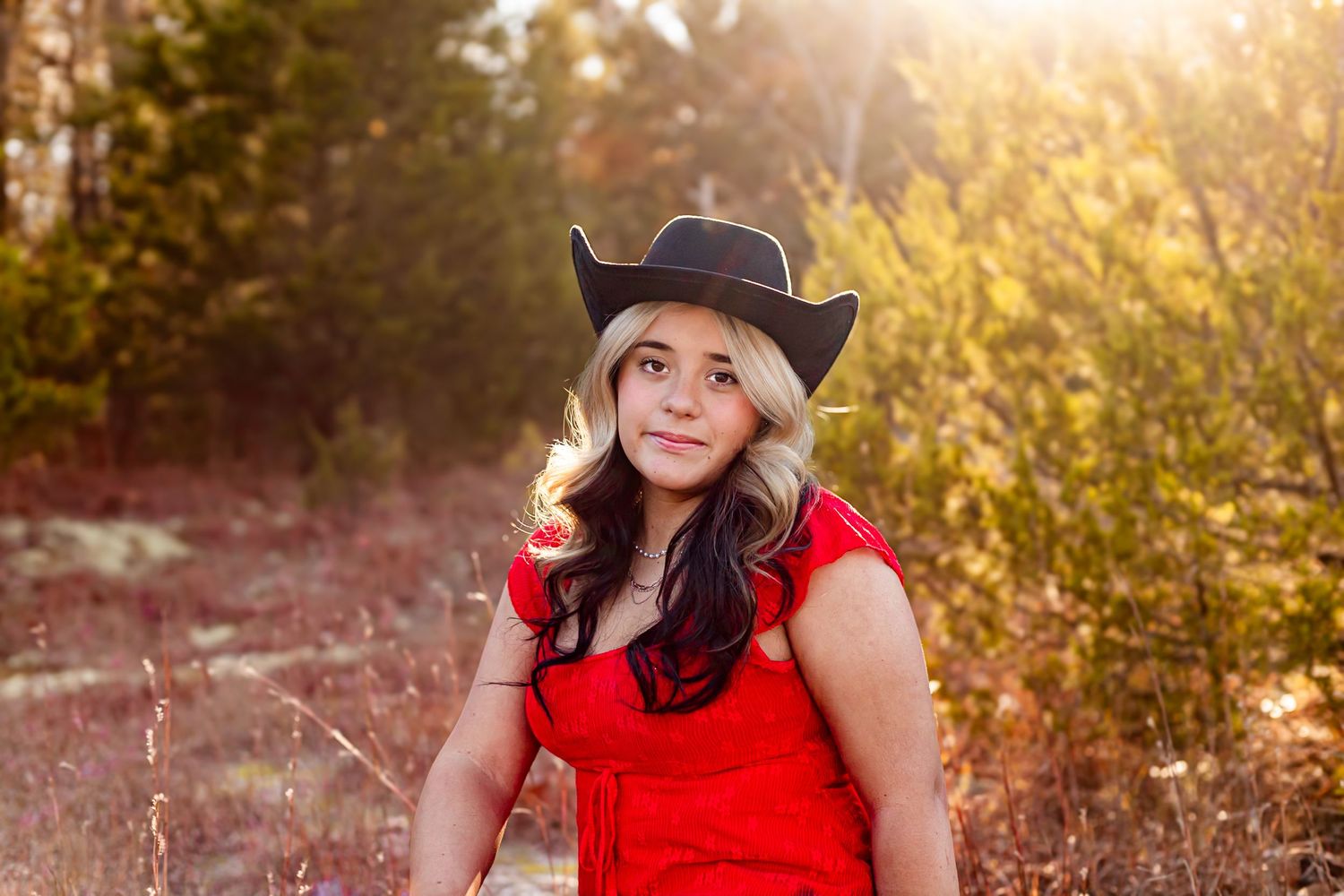 Young woman in red dress and black cowboy hat posing outdoors in golden sunlight with autumn foliage background.