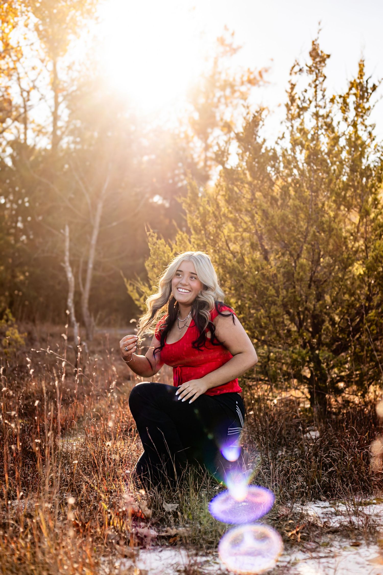 Smiling blonde woman in red top and black pants crouching in sunlit autumn field with trees behind her.