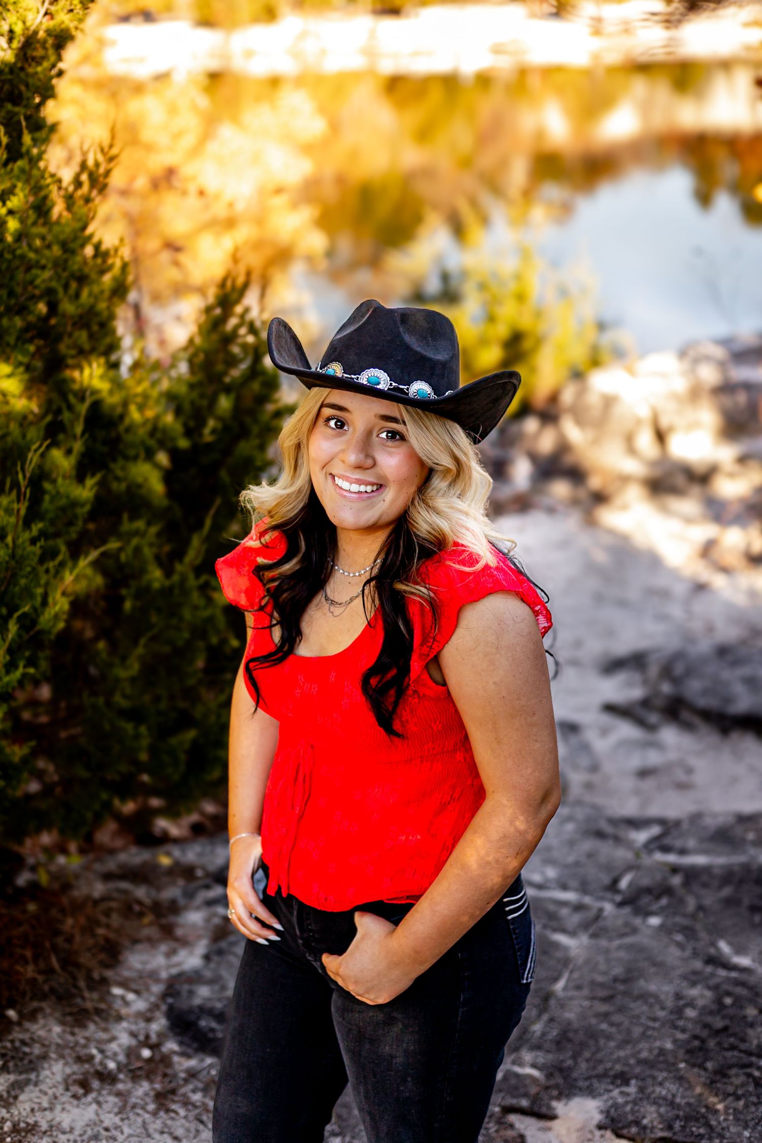 Smiling blonde woman wearing black cowboy hat and red top standing outdoors in warm golden natural light.