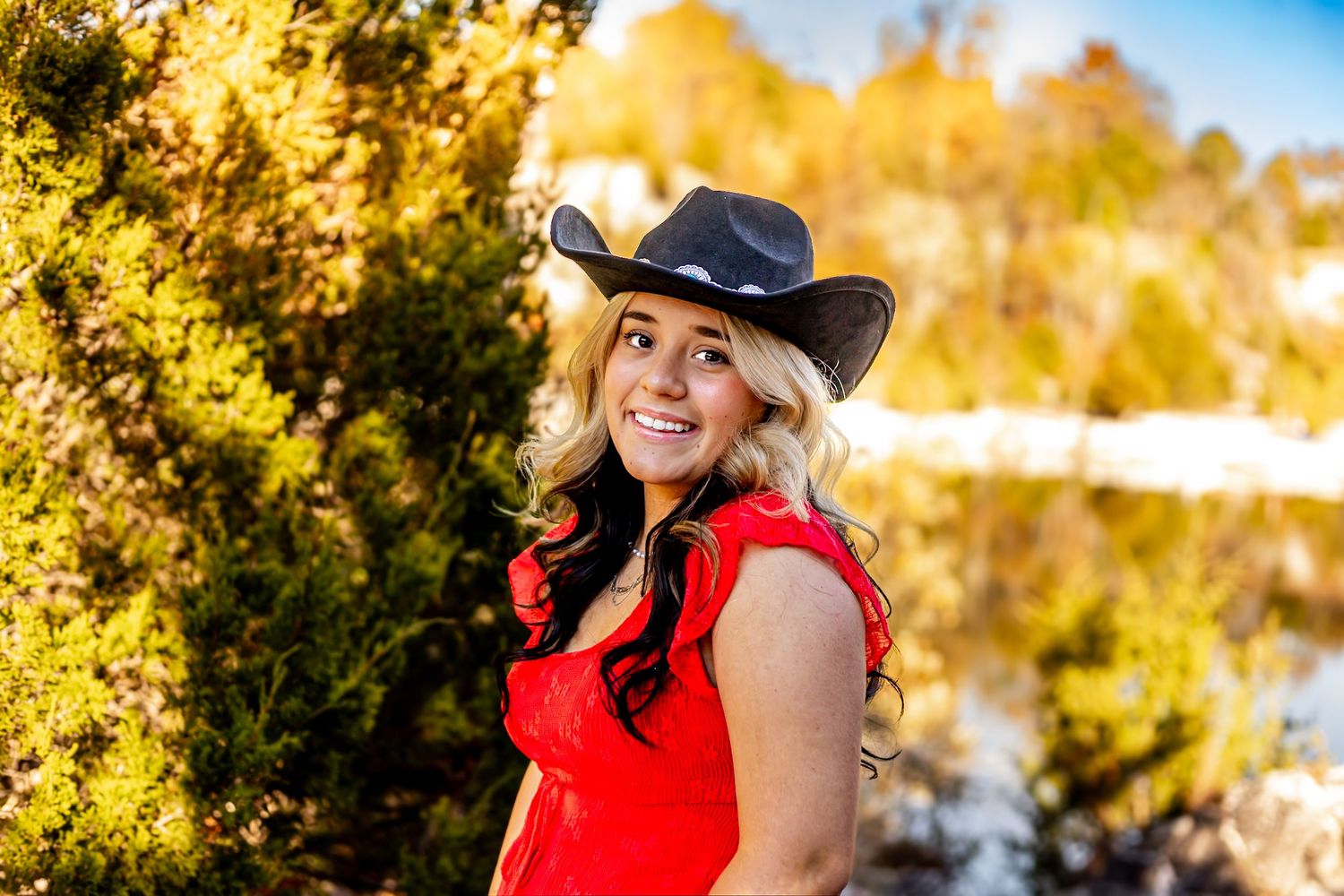 Smiling blonde woman in red dress and black cowboy hat posing outdoors among golden autumn trees.