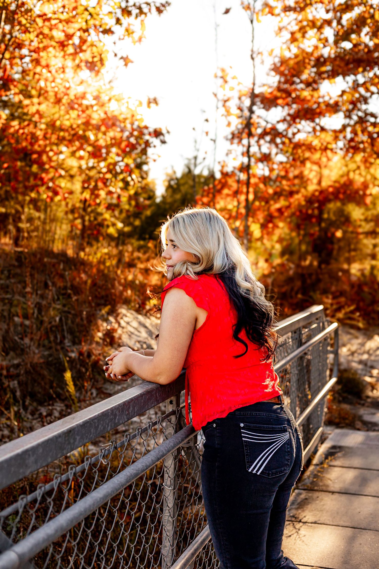 Woman in red top and jeans leans on bridge railing surrounded by vibrant autumn foliage in golden light.