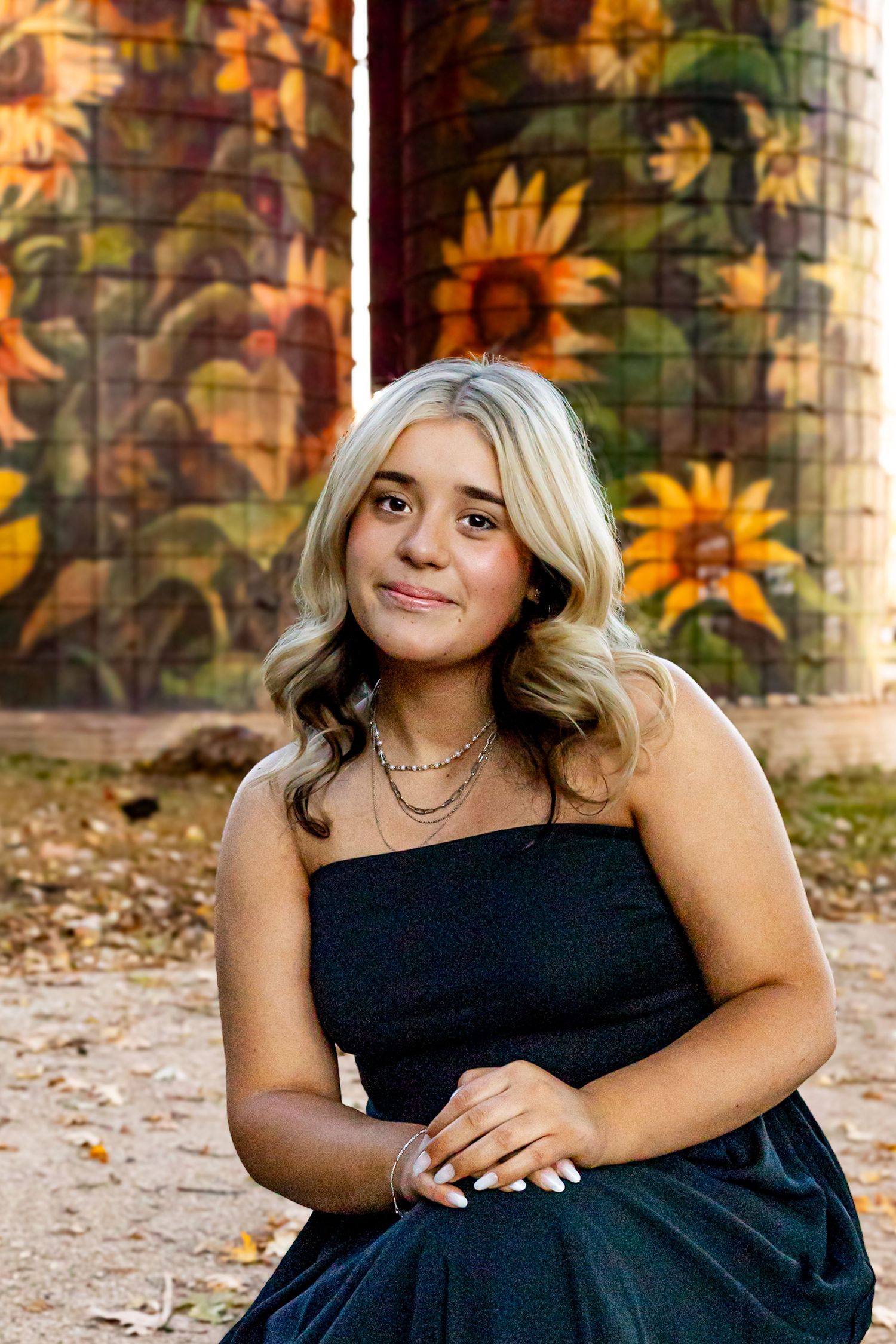 Young blonde woman in black strapless dress poses in front of colorful sunflower mural outdoors.