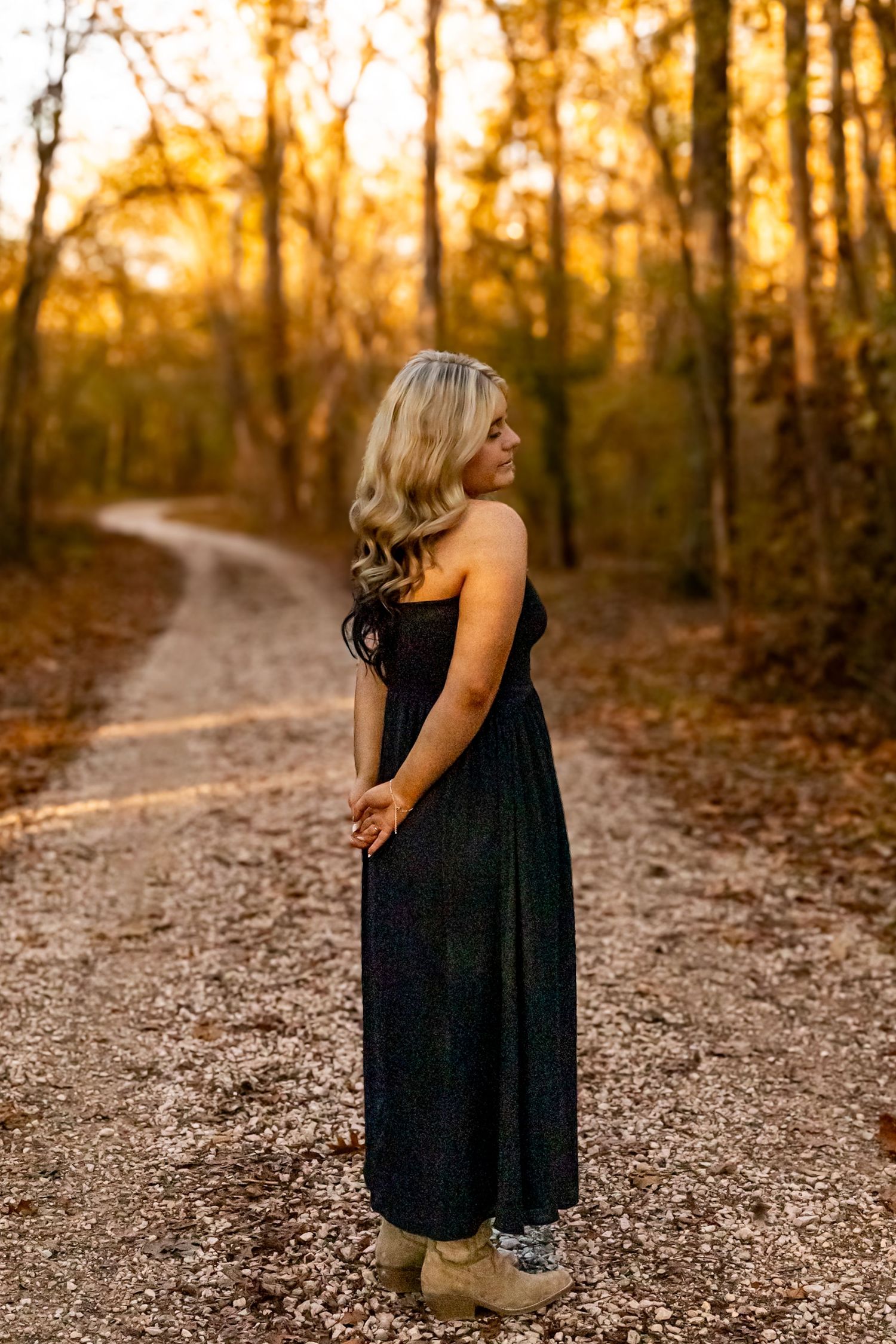 Young blonde woman in black maxi dress and boots standing on a winding forest path during golden hour autumn sunset.
