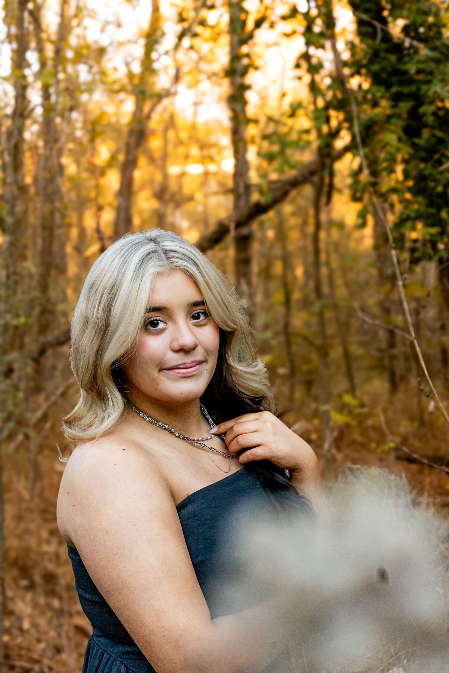 Young blonde woman in a navy strapless dress poses gracefully in a sunlit autumn forest setting.