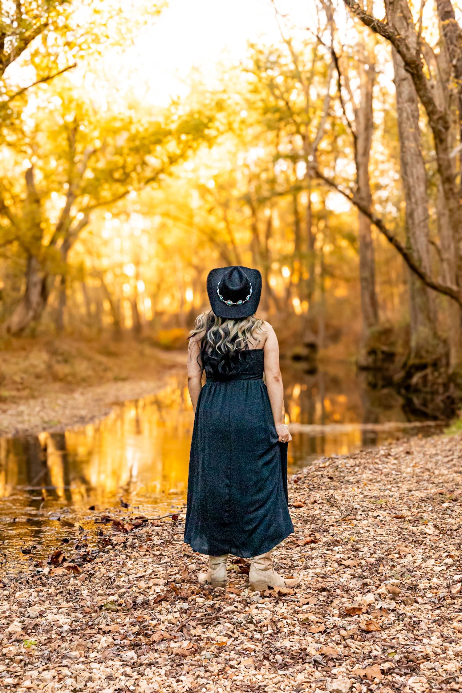 Woman in teal maxi dress and black cowboy hat stands by a reflective creek in golden autumn forest.