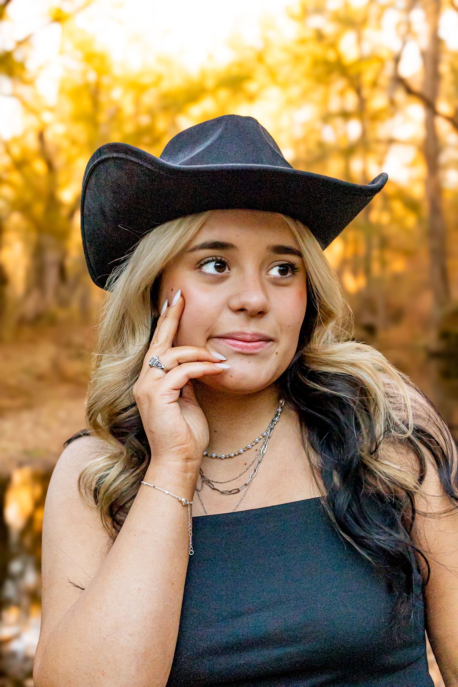 Young blonde woman in black cowboy hat and top posing thoughtfully outdoors during golden autumn foliage season.