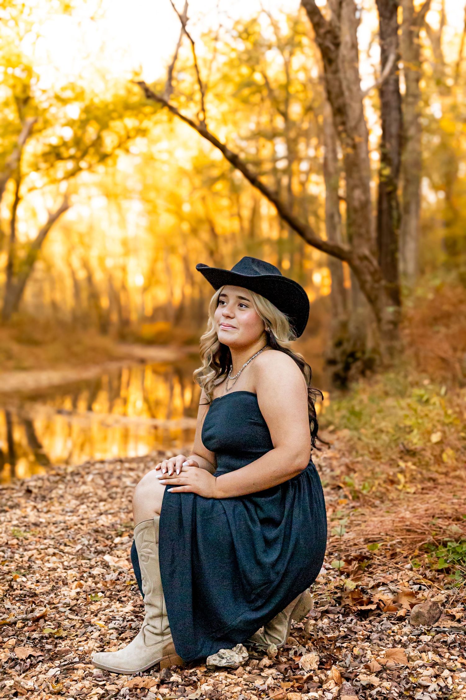 Woman in black cowboy hat and navy dress crouches among autumn leaves by a golden forest pond.