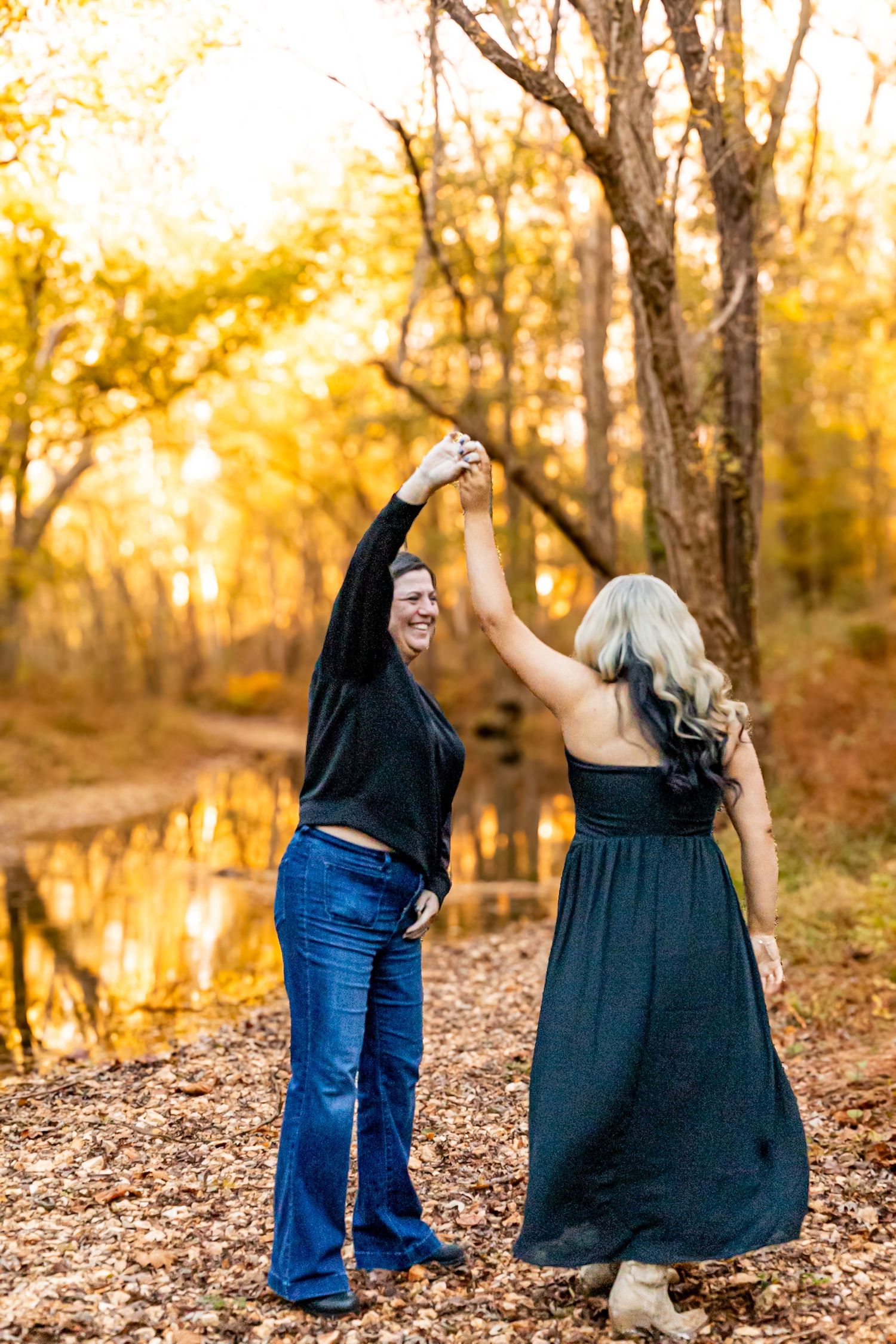 Two women dancing together by a creek surrounded by golden fall foliage in an autumn forest.