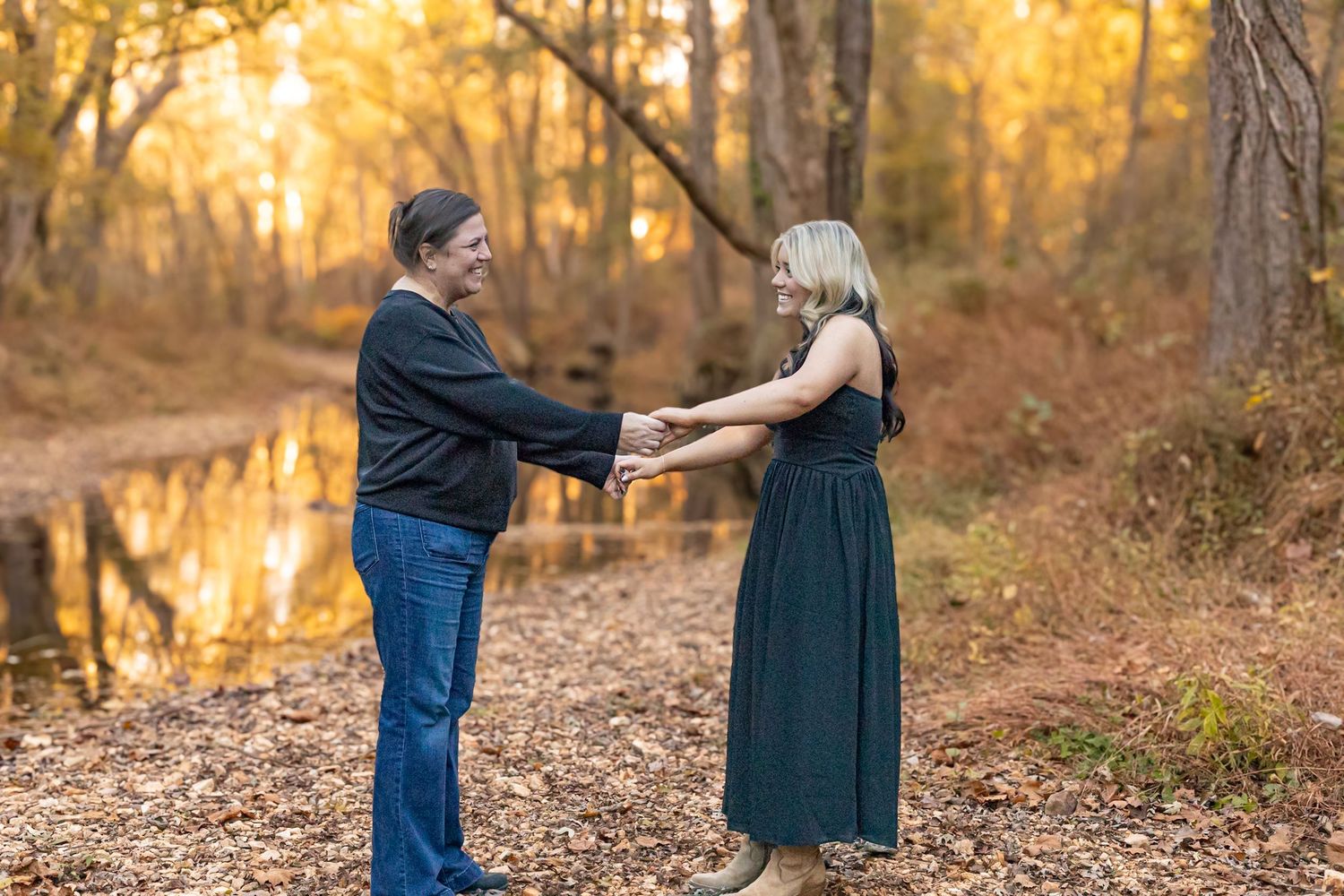 Couple holding hands and dancing together in an autumn forest with golden foliage and a reflective pond backdrop.