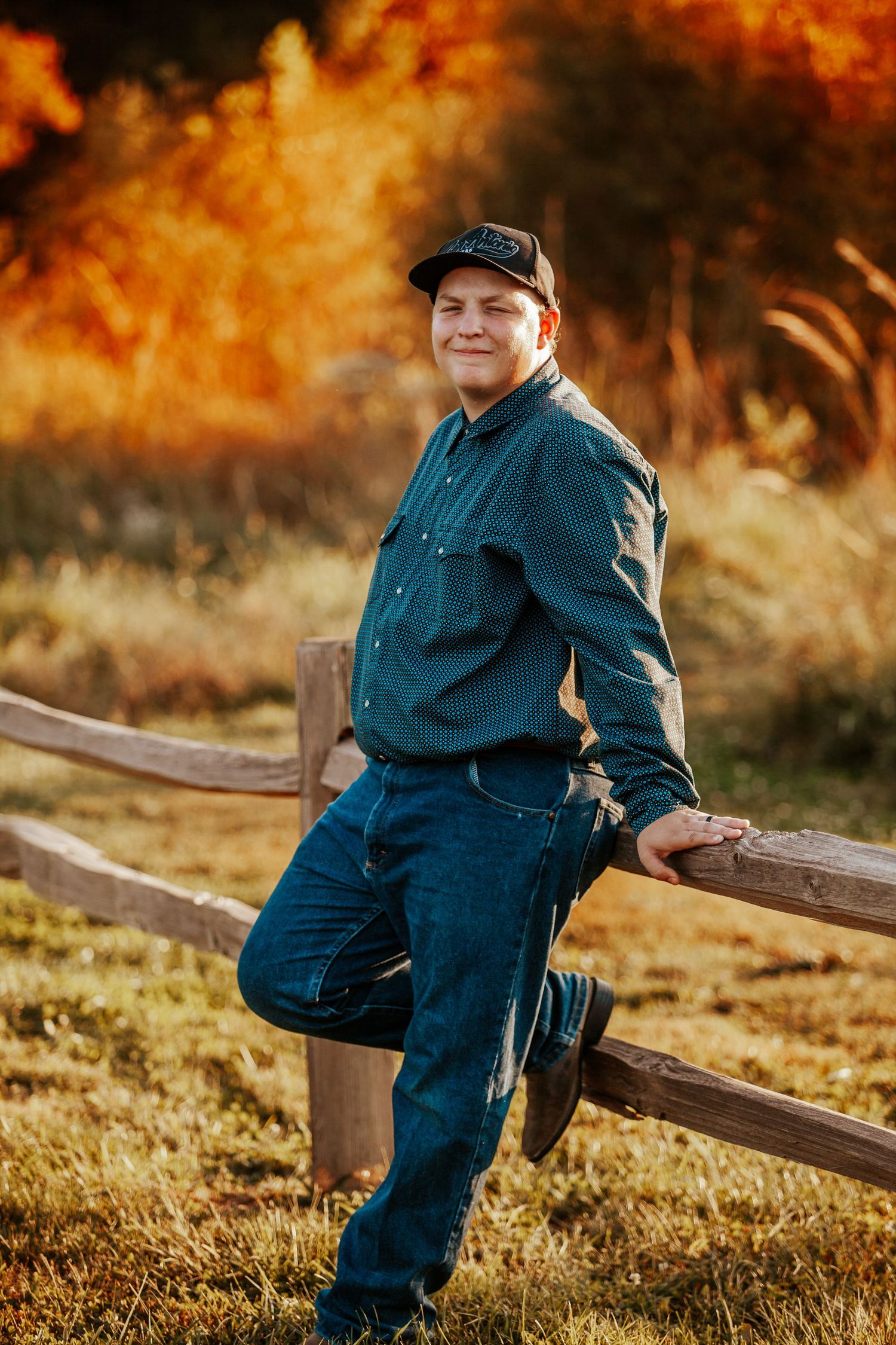 Man in teal shirt and cap leaning on wooden fence in autumn field with golden foliage background.