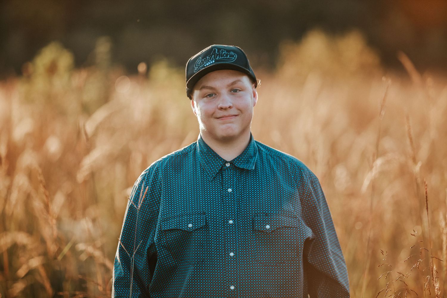 Young man in teal shirt and cap smiling in golden wheat field during sunset.
