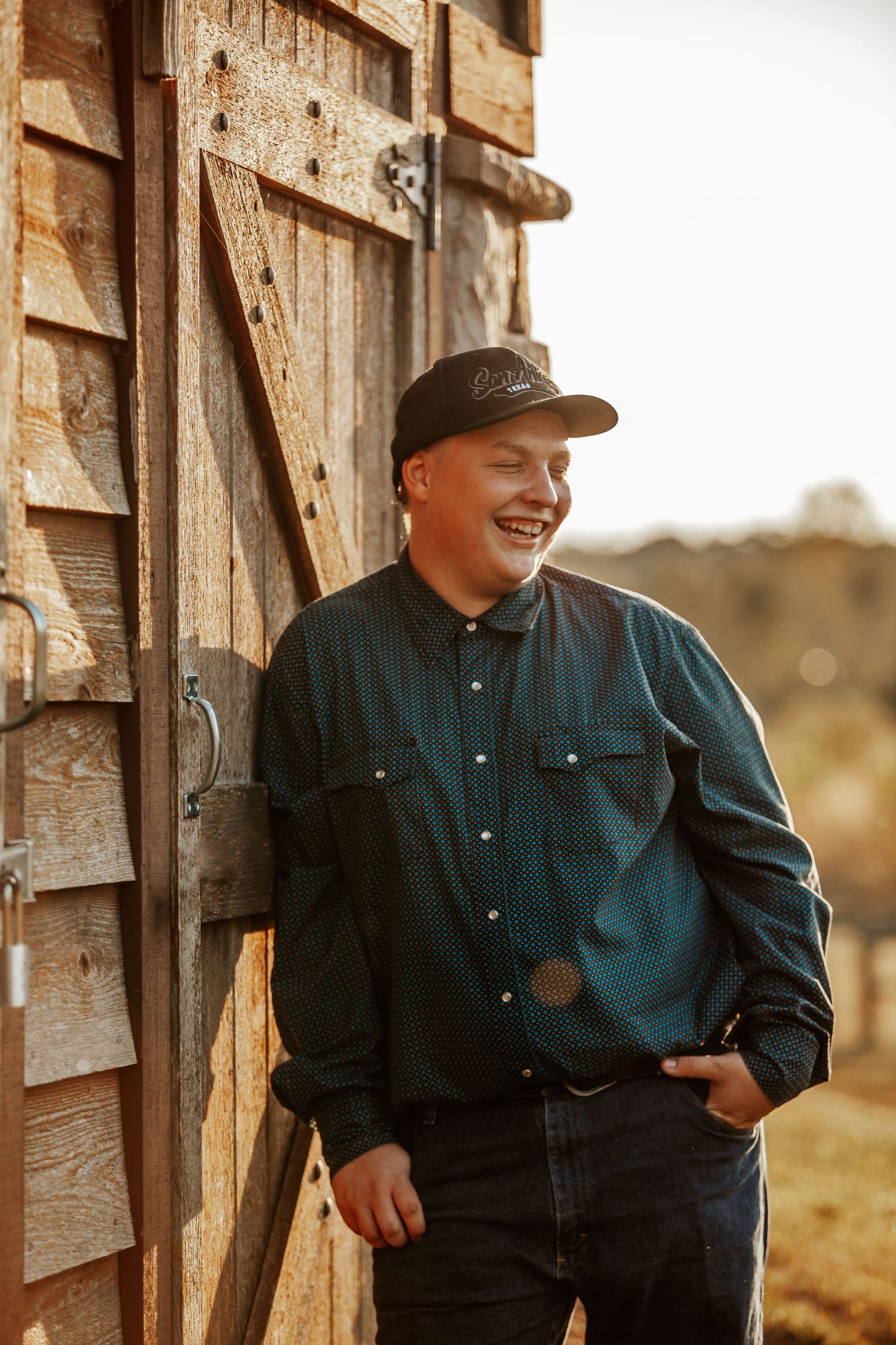 Smiling young man in a black cap and teal shirt leaning against a rustic wooden barn door outdoors.