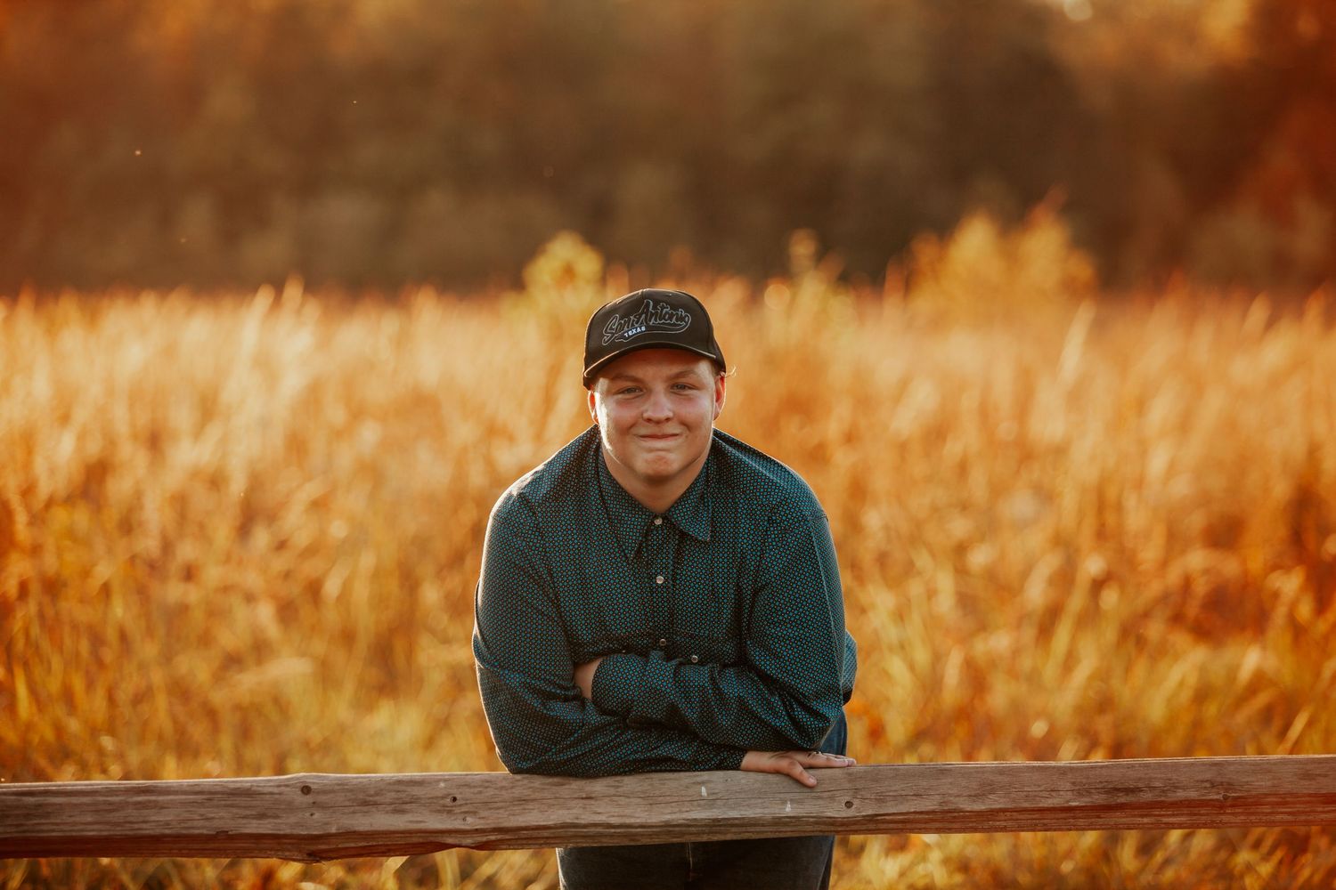 Smiling man in green jacket and cap leaning on wooden fence in golden autumn field at sunset.
