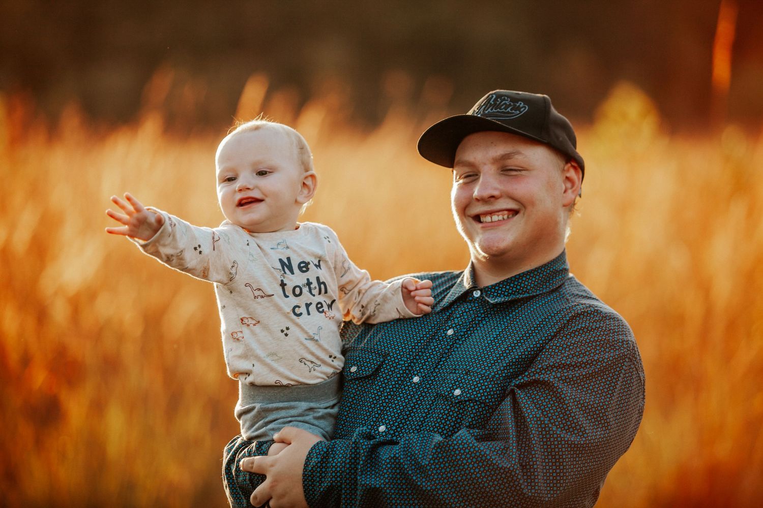 A smiling young man holds a happy baby in a sunlit golden field, both enjoying a warm outdoor moment together.