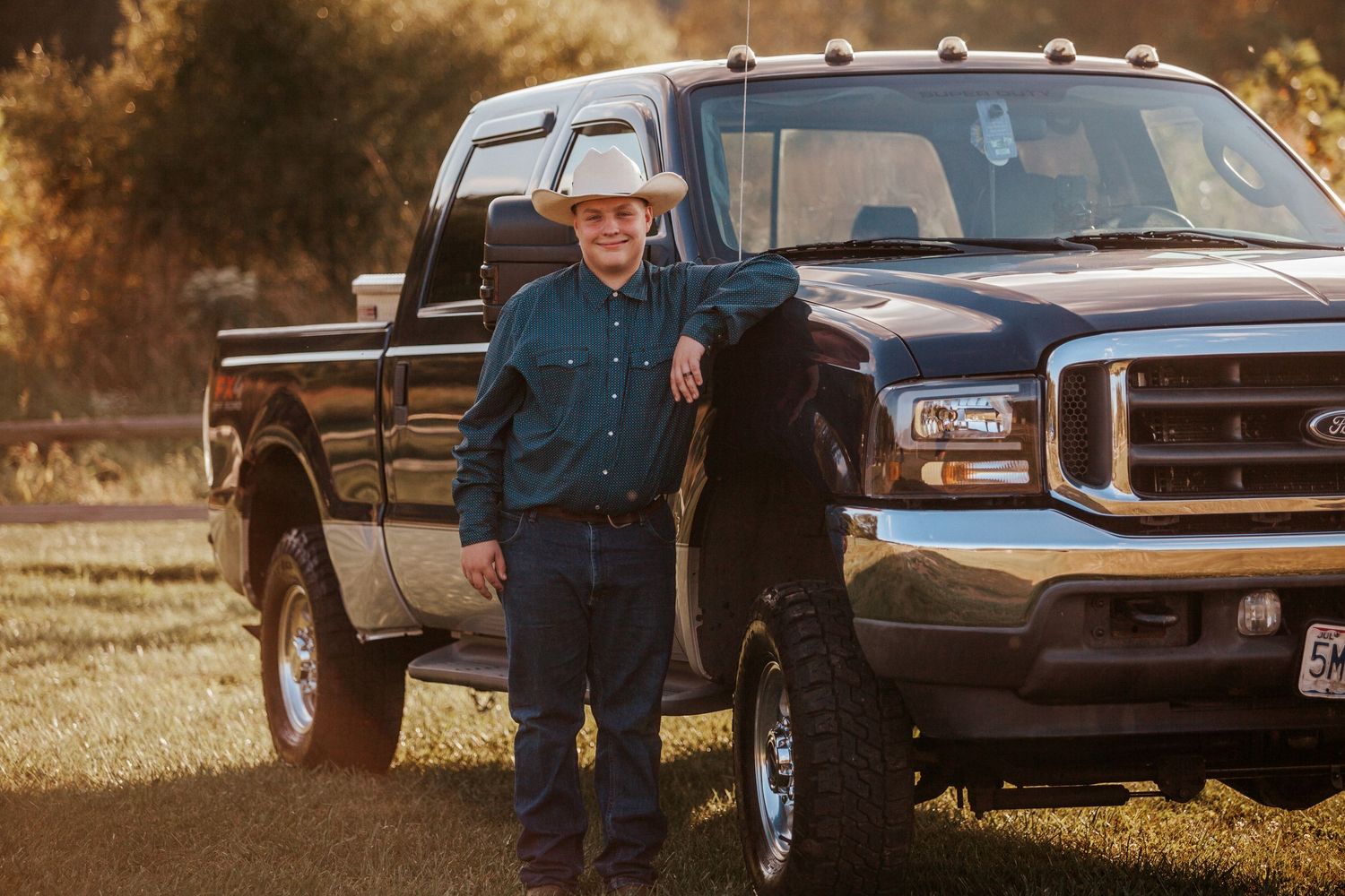 Man in cowboy hat leaning against black Ford pickup truck in golden hour sunlight on a rural farm.