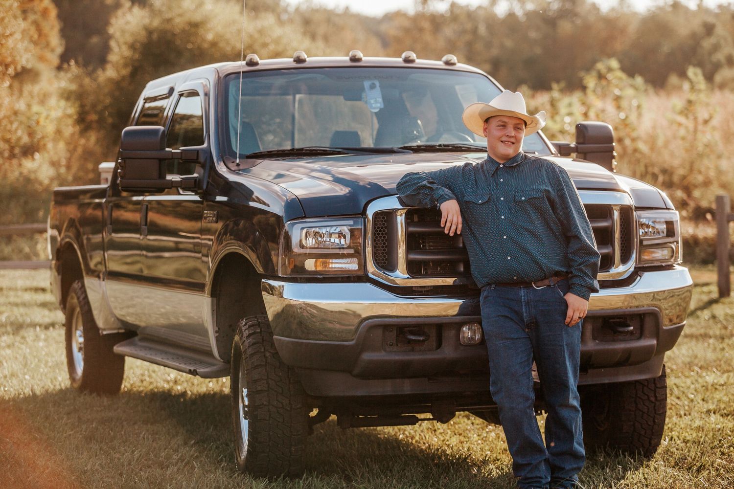 Man in cowboy hat leaning against black Ford pickup truck in rural countryside setting.