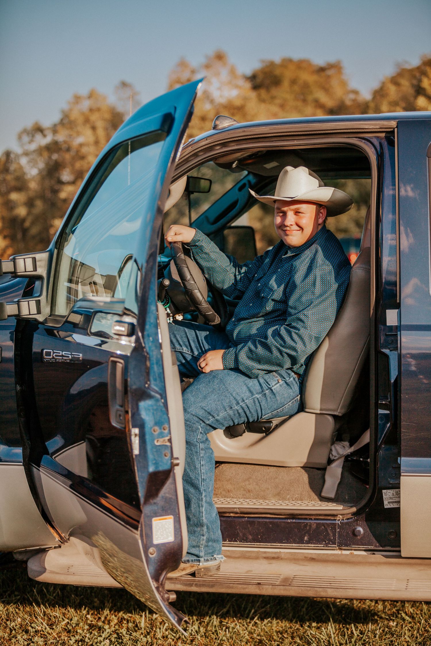 Man in white cowboy hat and denim outfit sitting in truck cab with door open, smiling outdoors.