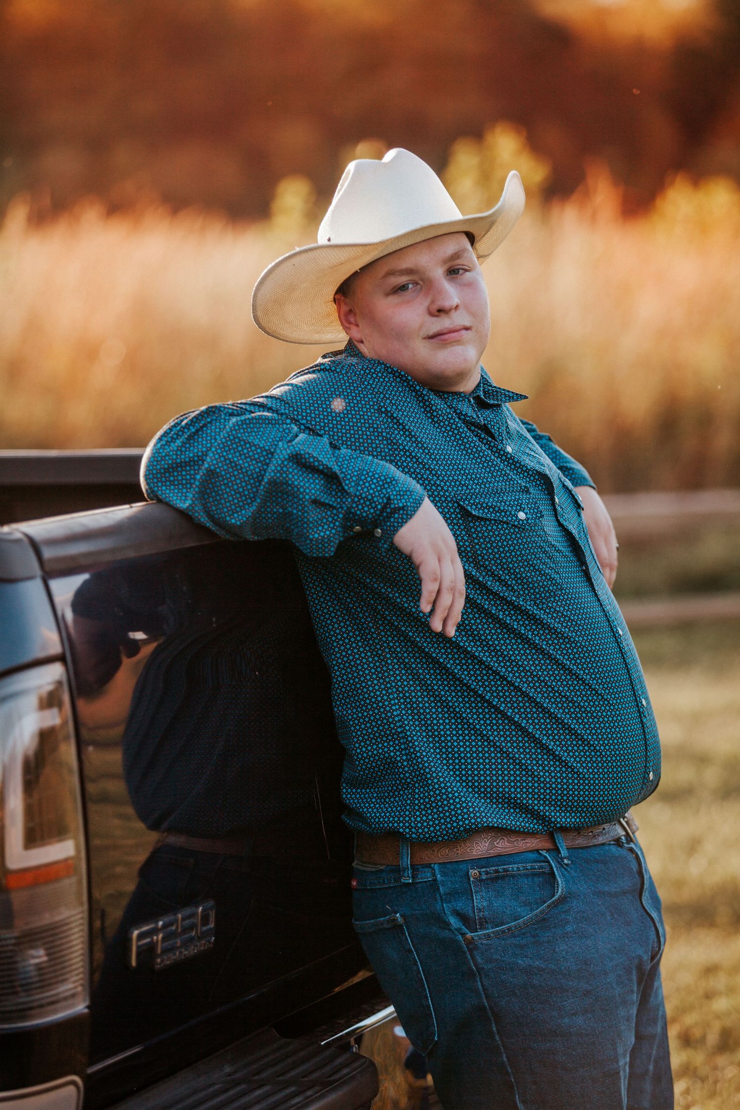 Young man in white cowboy hat and teal shirt leaning against a Ford truck in a golden field at sunset.