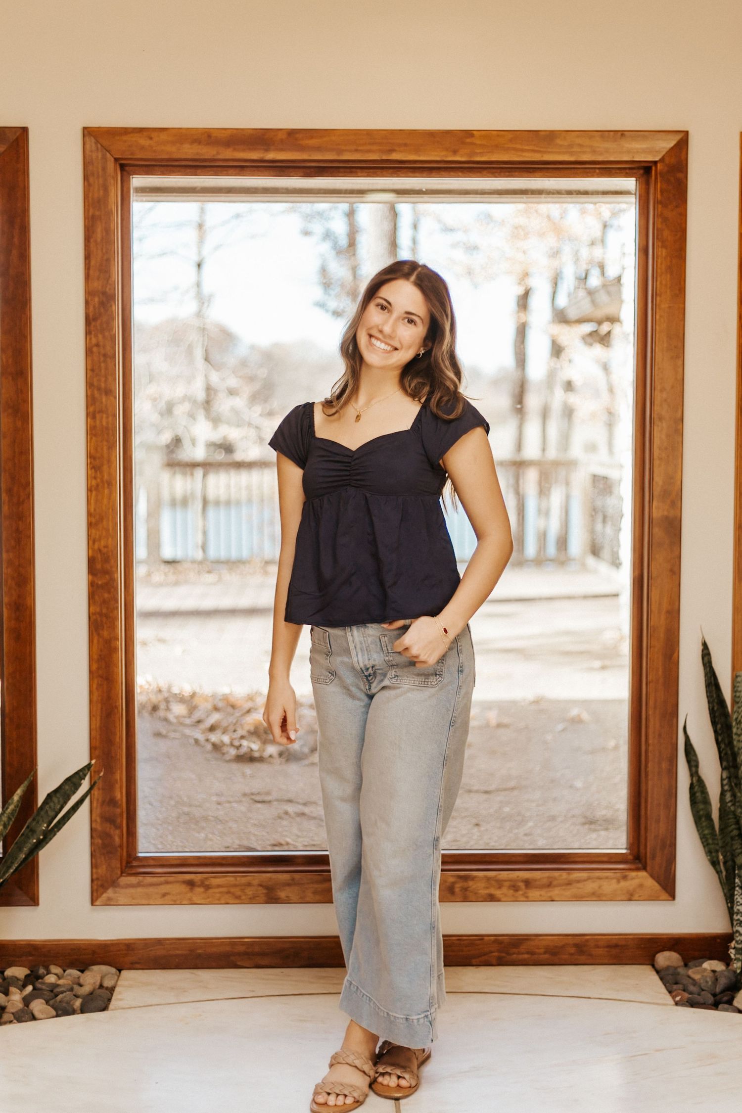 Woman in black babydoll top and wide-leg jeans smiling in front of large wooden-framed window.