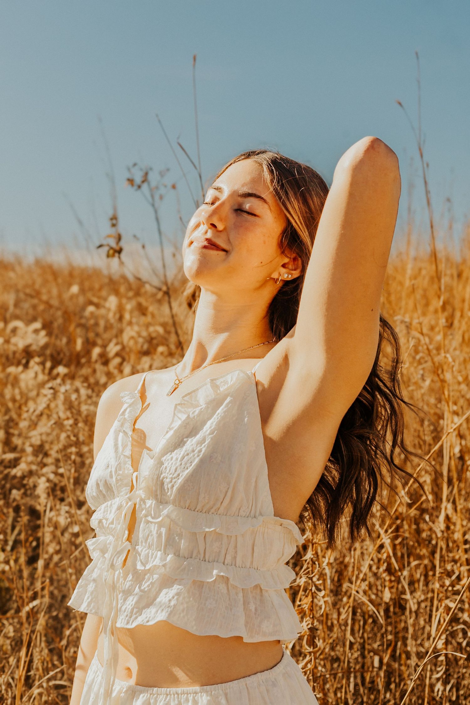 Young woman in white ruffled crop top posing in golden wheat field under blue sky.