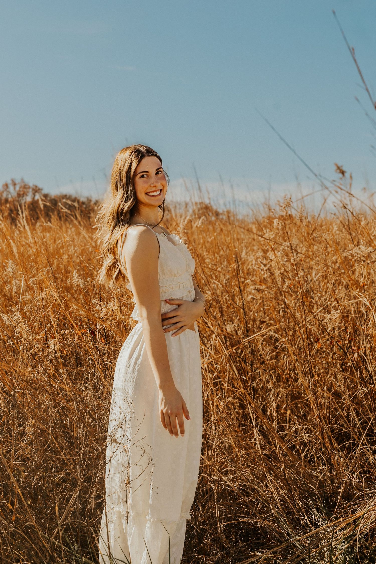 Young woman in white lace dress smiling in golden wheat field under blue sky.