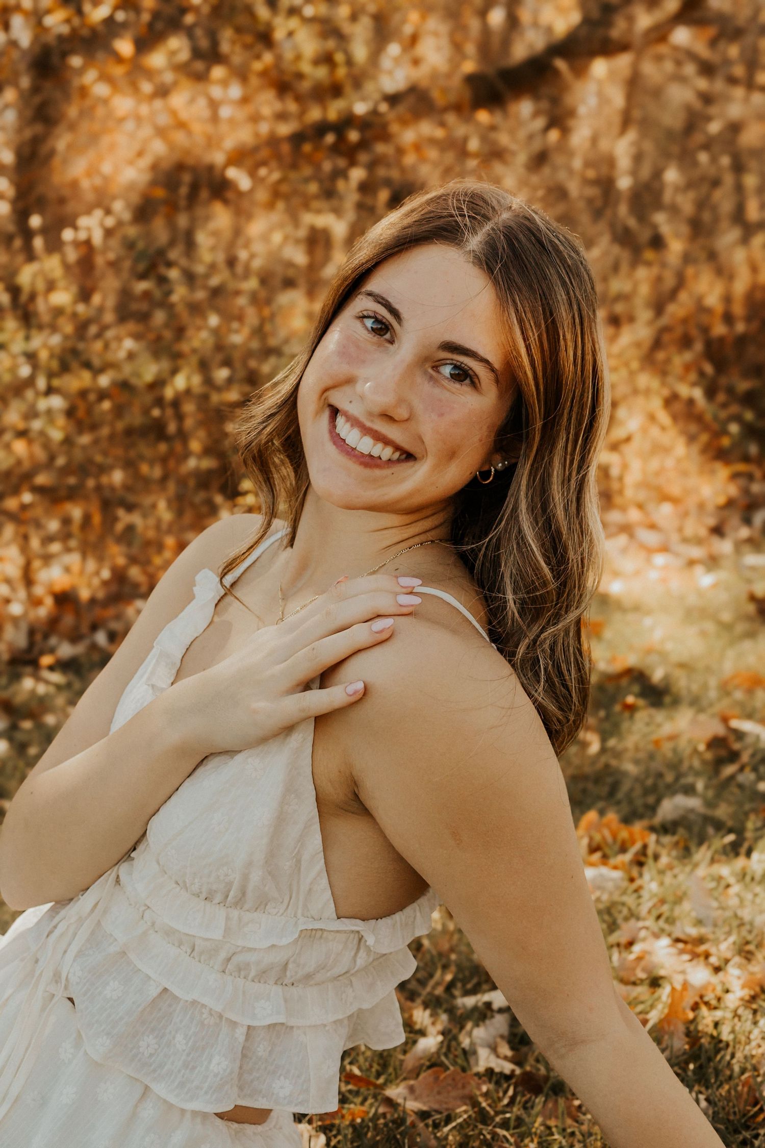 Smiling young woman in white ruffled dress posing among autumn foliage.
