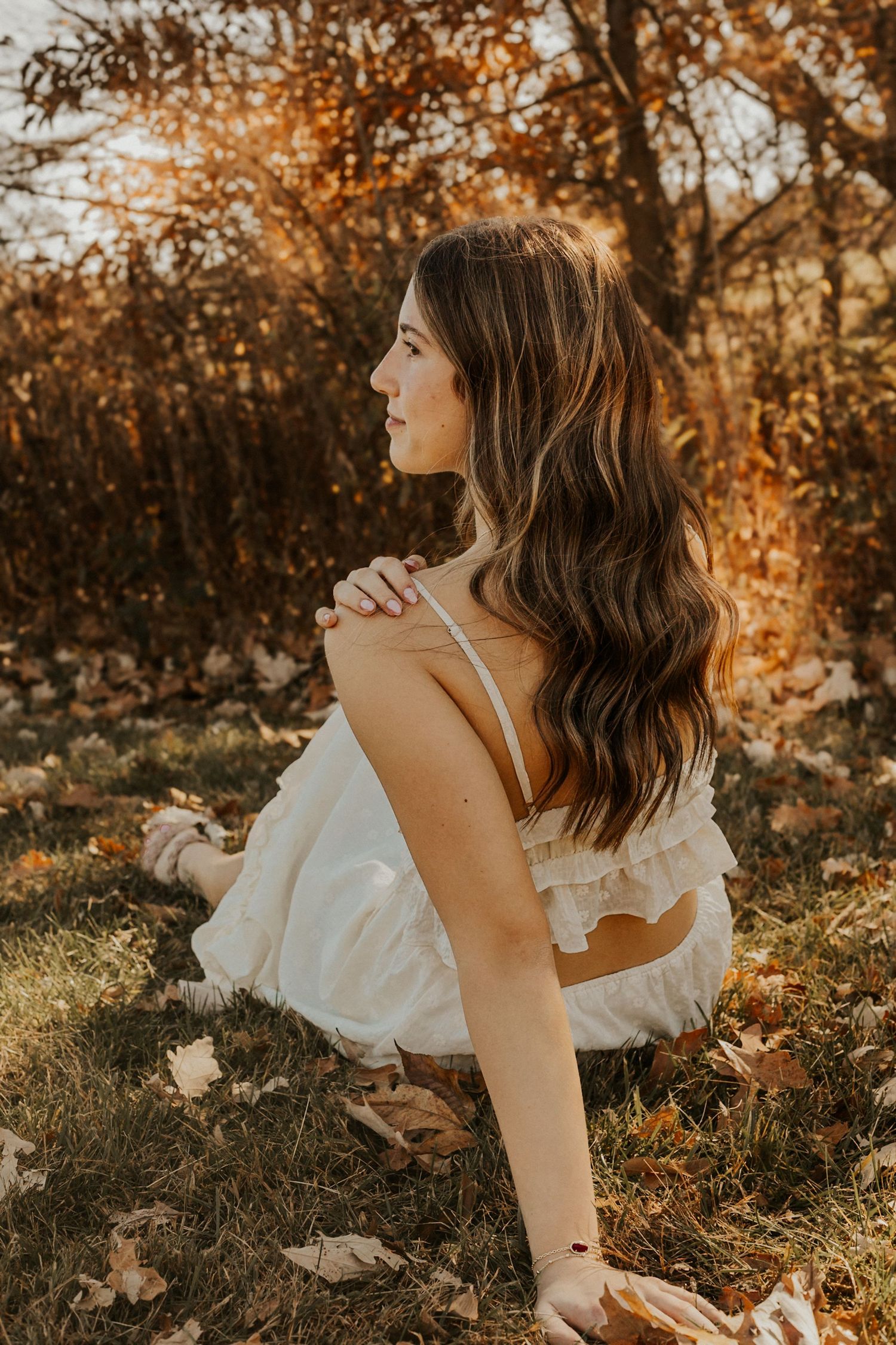 Young woman in white dress sitting on autumn leaves, golden hour sunlight illuminating her long wavy brown hair.