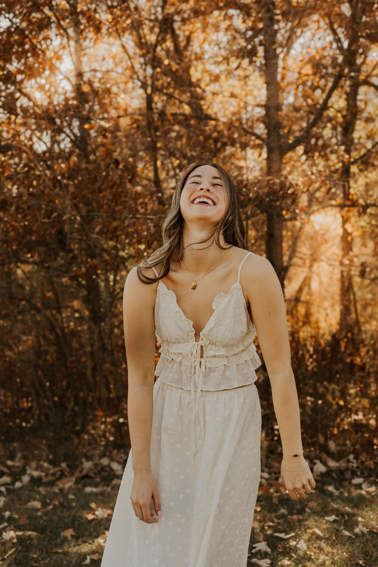 Joyful young woman laughing in white lace dress surrounded by fall foliage in warm golden sunlight.