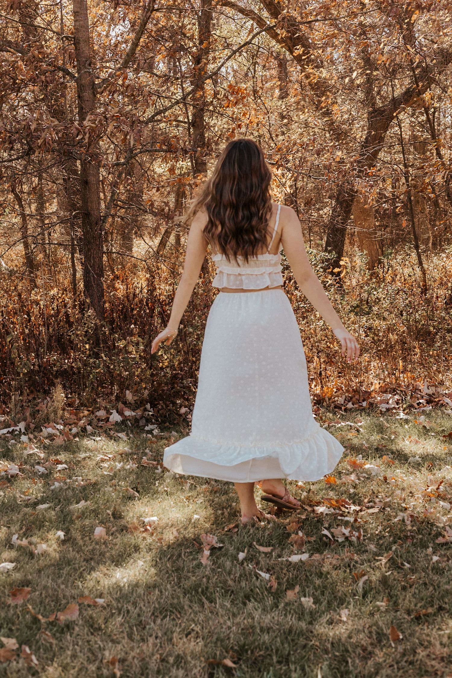 Woman in white ruffled maxi dress walking through sunlit autumn woodland from behind.