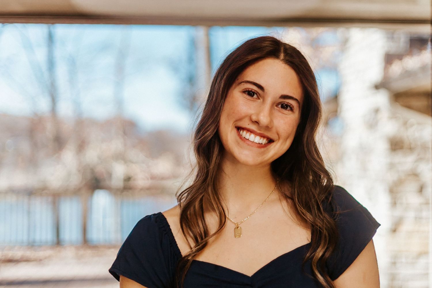 Smiling young woman with long brown hair wearing a navy off-shoulder top and gold necklace outdoors.