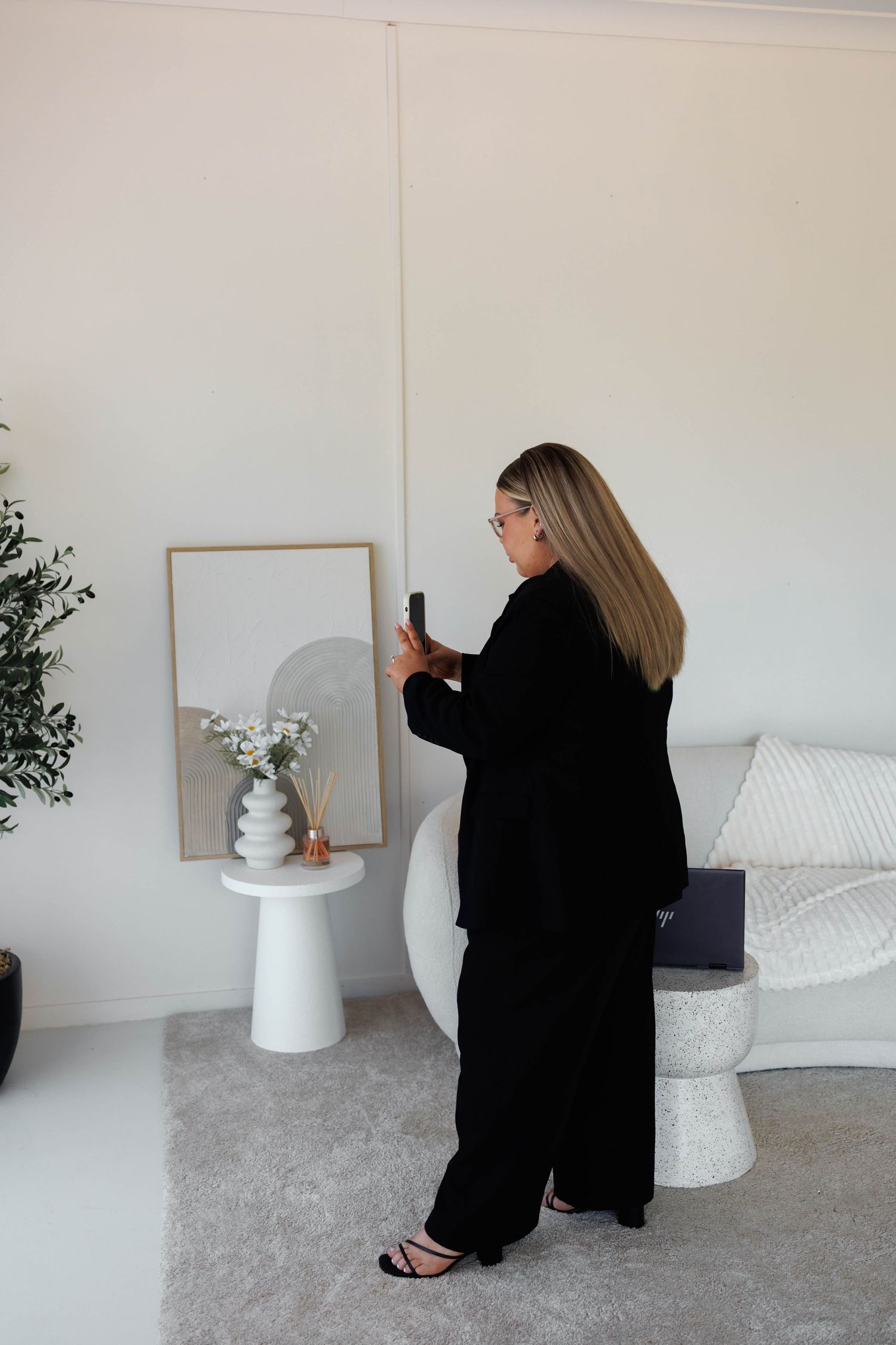 Woman in black suit photographing minimalist white living room decor with smartphone, wearing heels and glasses.