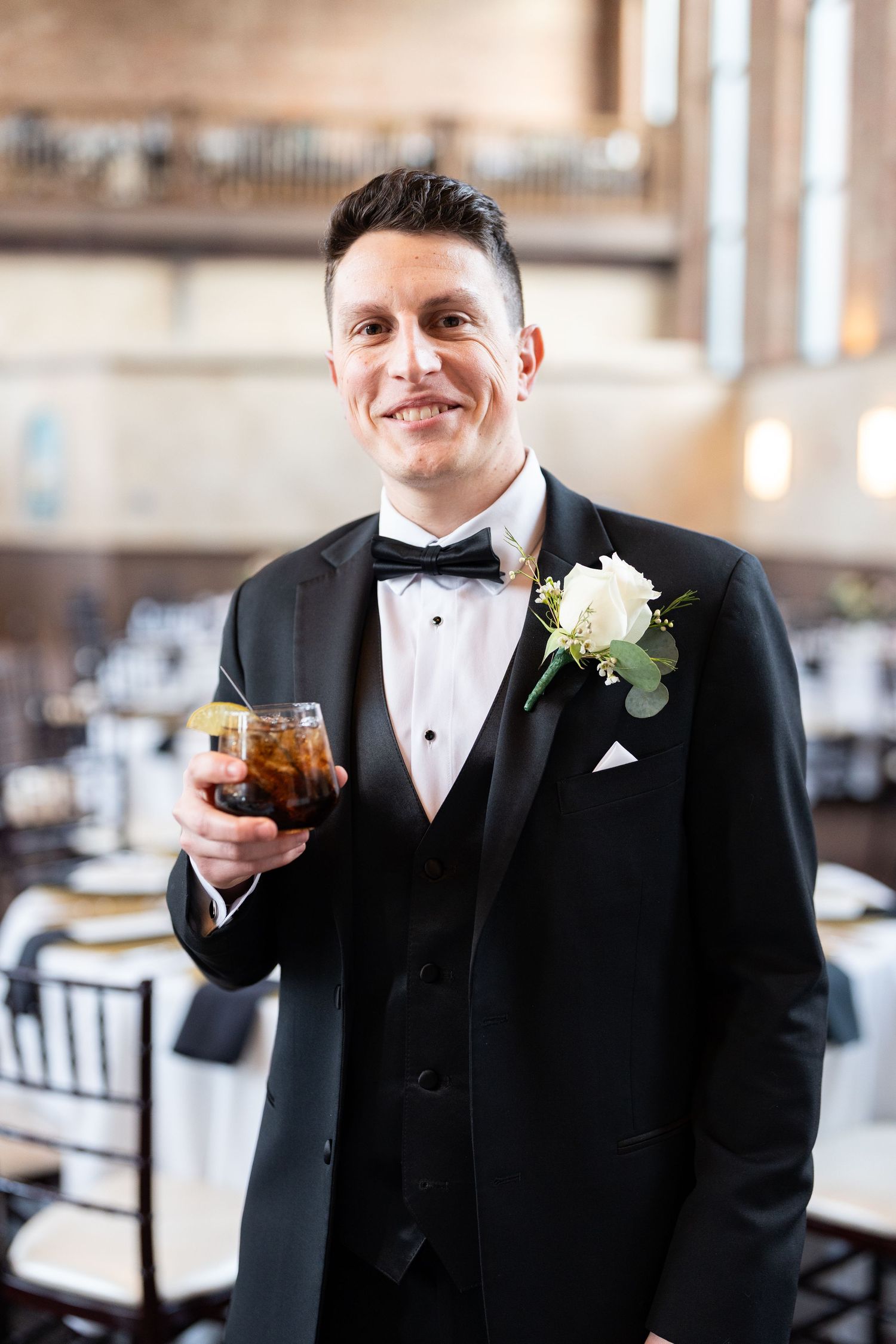 Smiling groom in black tuxedo with white boutonniere holding a cocktail glass at the Monastery Cincinatti wedding venue.