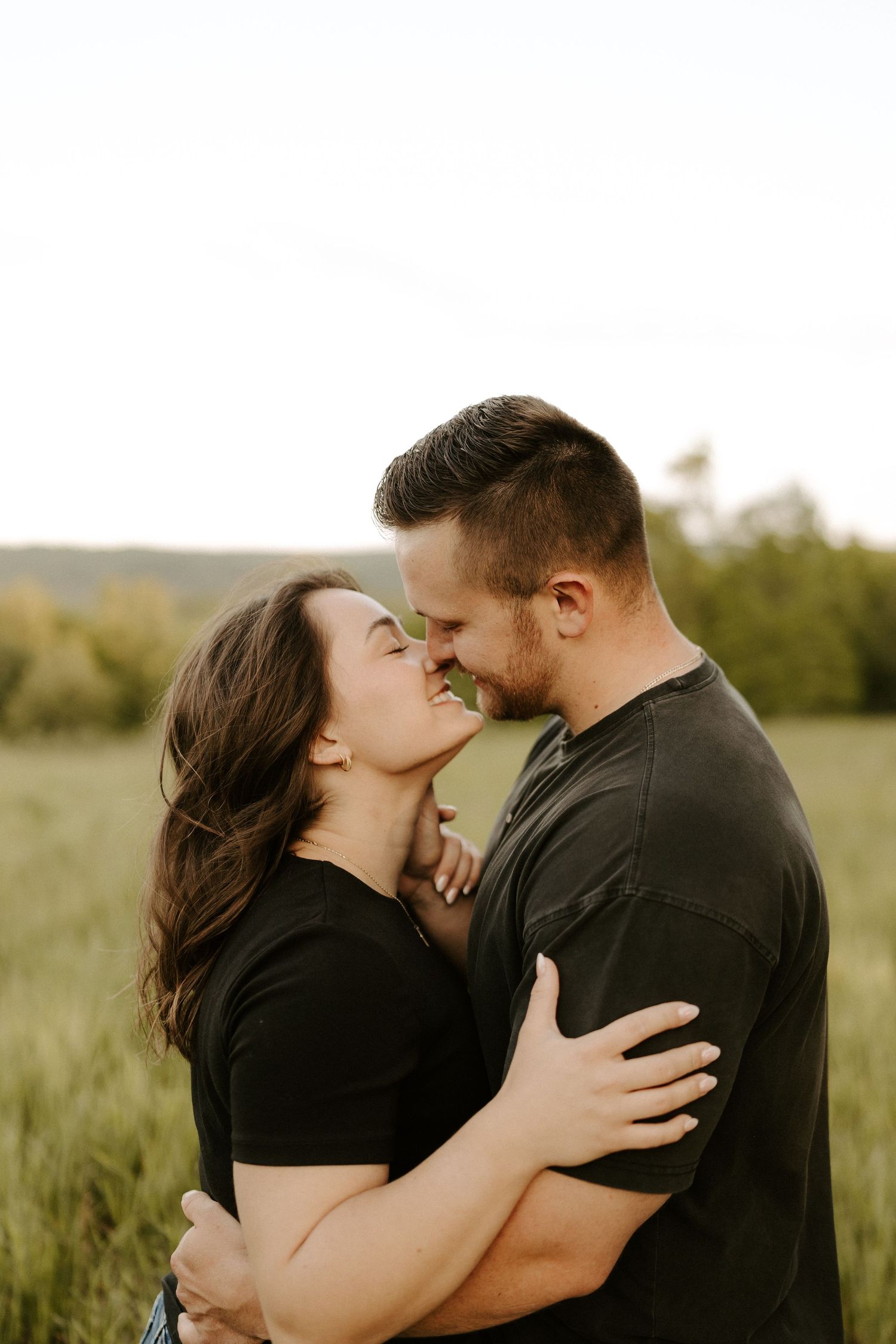 Couple sharing a tender almost-kiss while embracing in a lush green meadow at sunset.