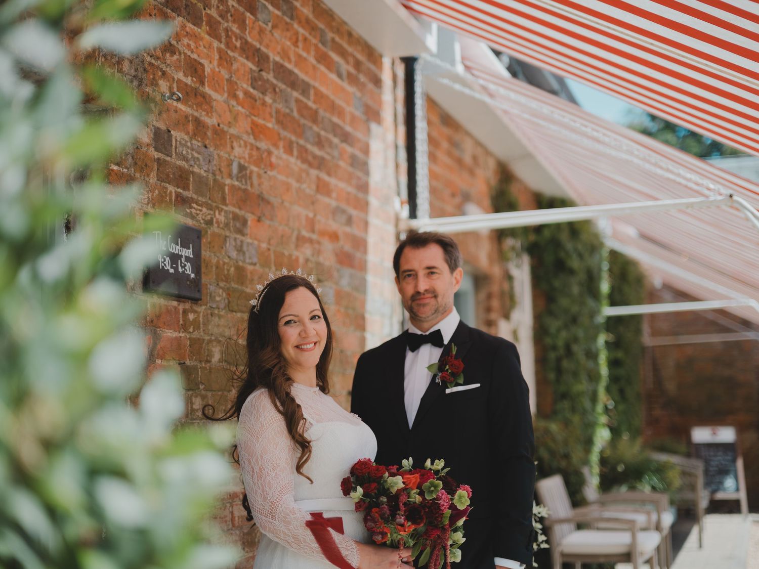 Bride and groom smiling together outside a brick venue with red striped awning, bride holding red bouquet.