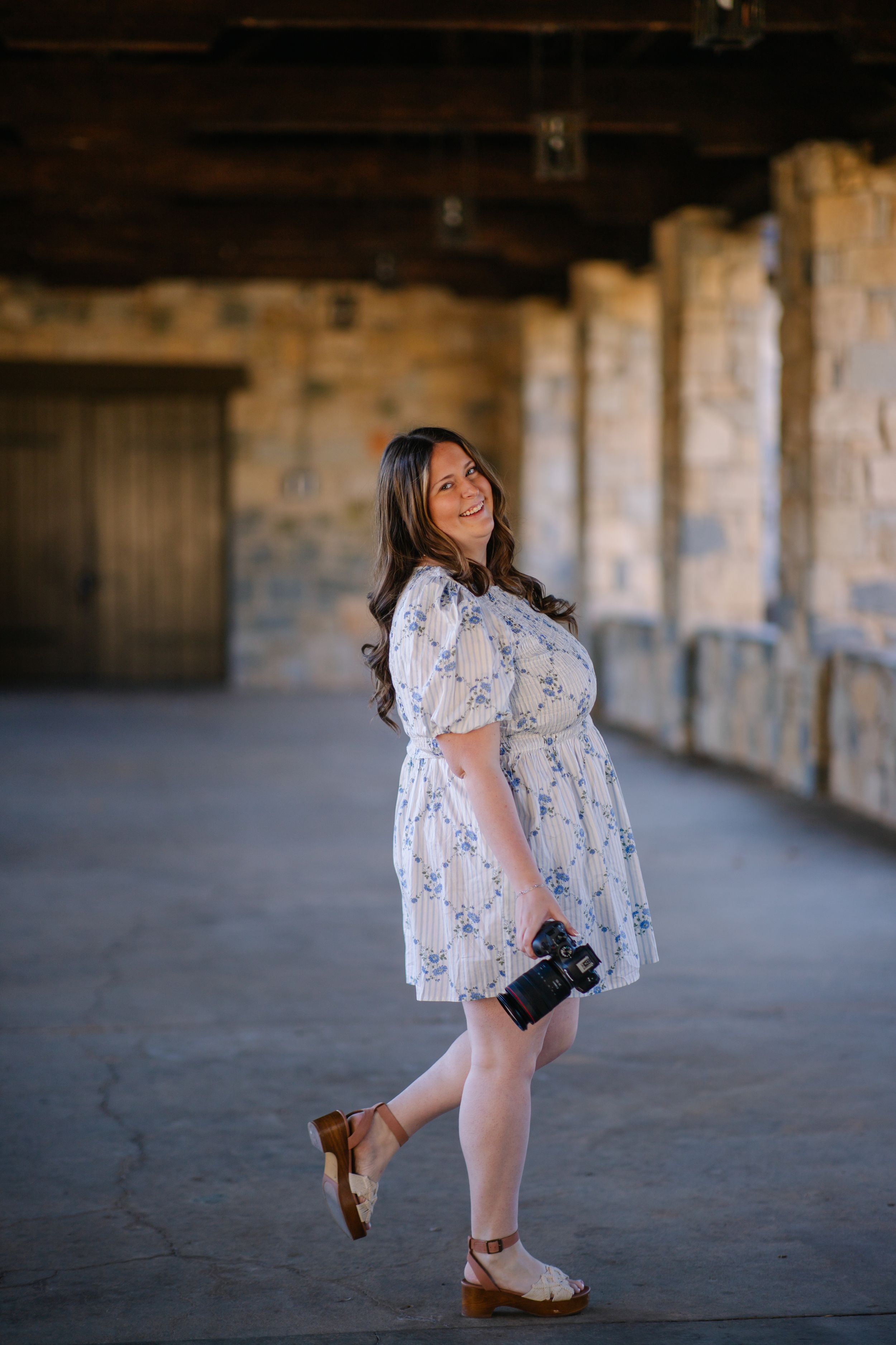 Smiling pregnant woman in floral dress holding camera, posing in rustic stone building with warm natural light.