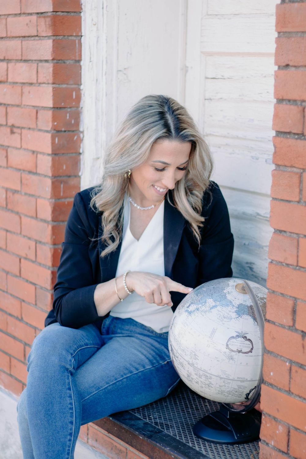 Smiling blonde woman in black blazer and jeans pointing at a vintage globe while sitting outdoors near a brick wall.