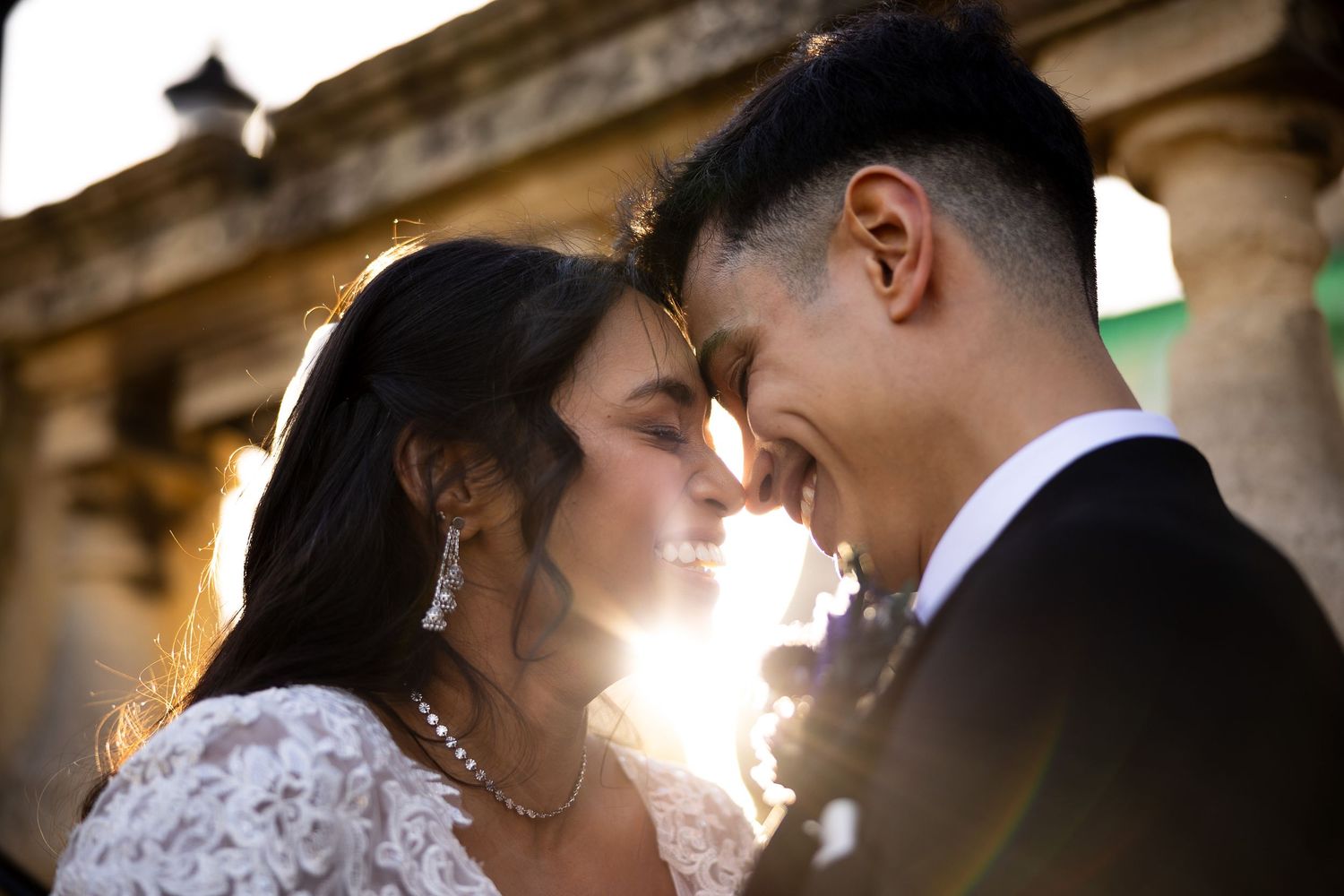 A bride and groom share an intimate forehead touch, backlit by golden sunset light at an outdoor wedding venue.