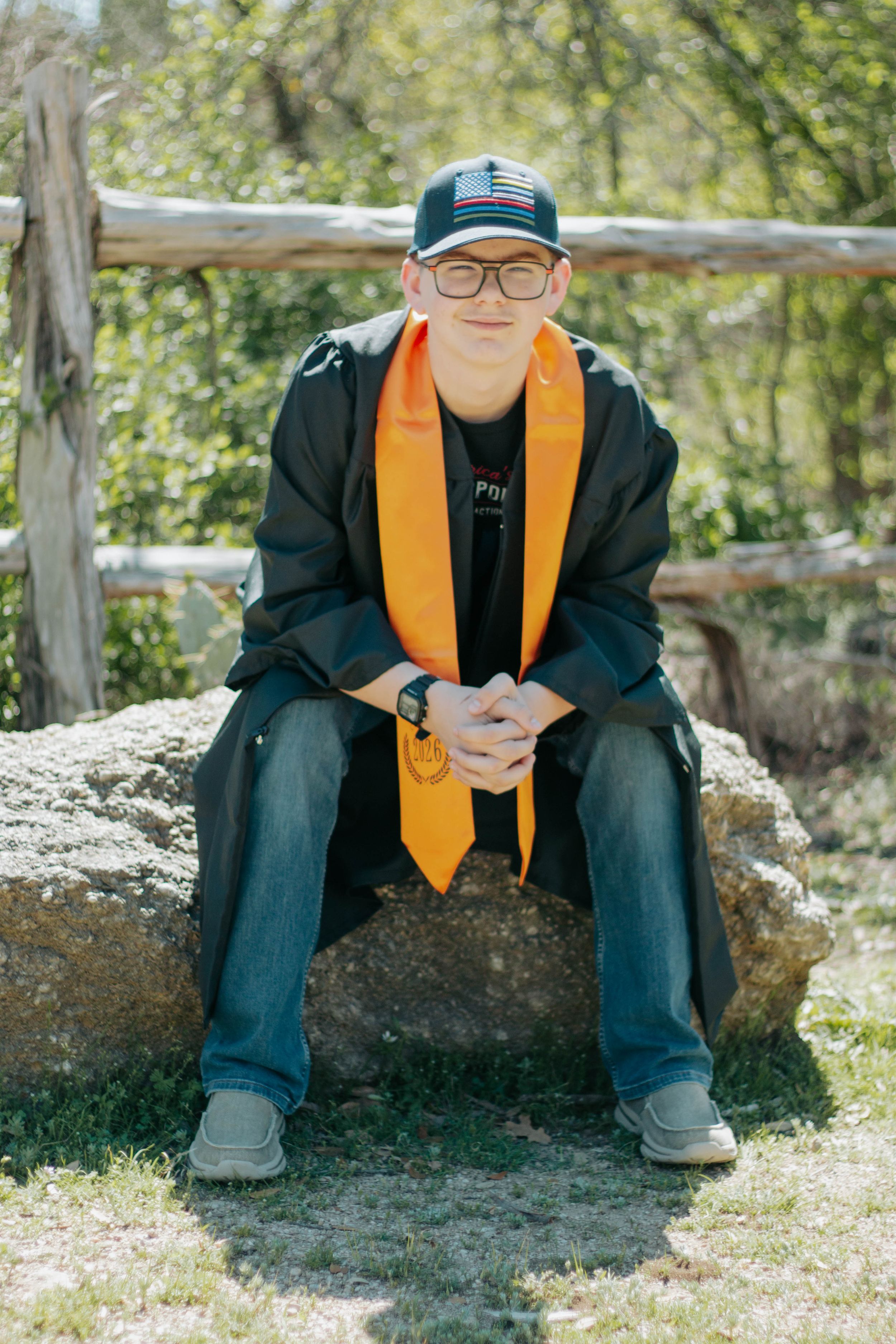 Young man in graduation stole, cap, and glasses sitting on a rock outdoors near a wooden fence.