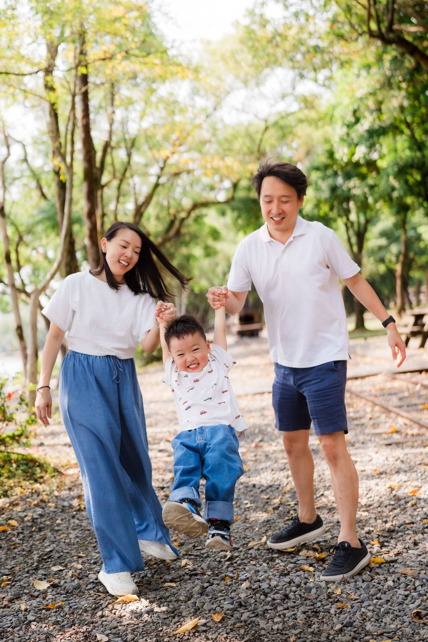 Happy Asian family with young toddler son swinging between parents outdoors in a sunny green park.