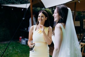Bride in white veil laughing with bridesmaid in yellow dress at outdoor wedding reception.
