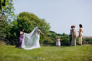 Wedding party helping bride with veil on green hillside lawn under clear blue sky.