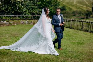 Bride in lace-trimmed veil and white gown walks with groom in navy suit on green hillside at outdoor wedding.