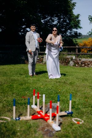 Bride in white gown plays ring toss lawn game with suited groom at outdoor wedding reception.