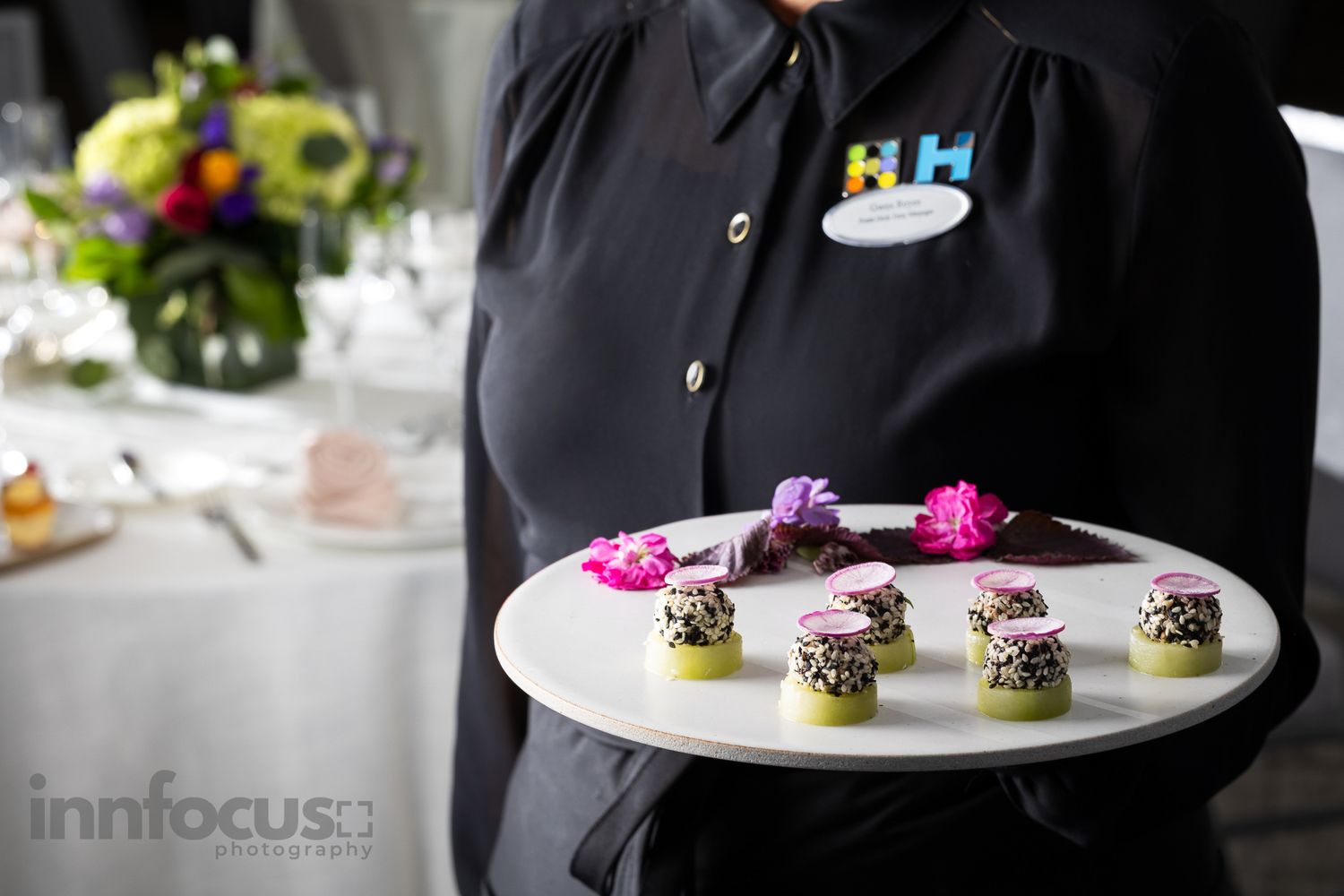 A catering server in black uniform holds a white plate of elegant bite-sized appetizers garnished with pink flowers at an event.