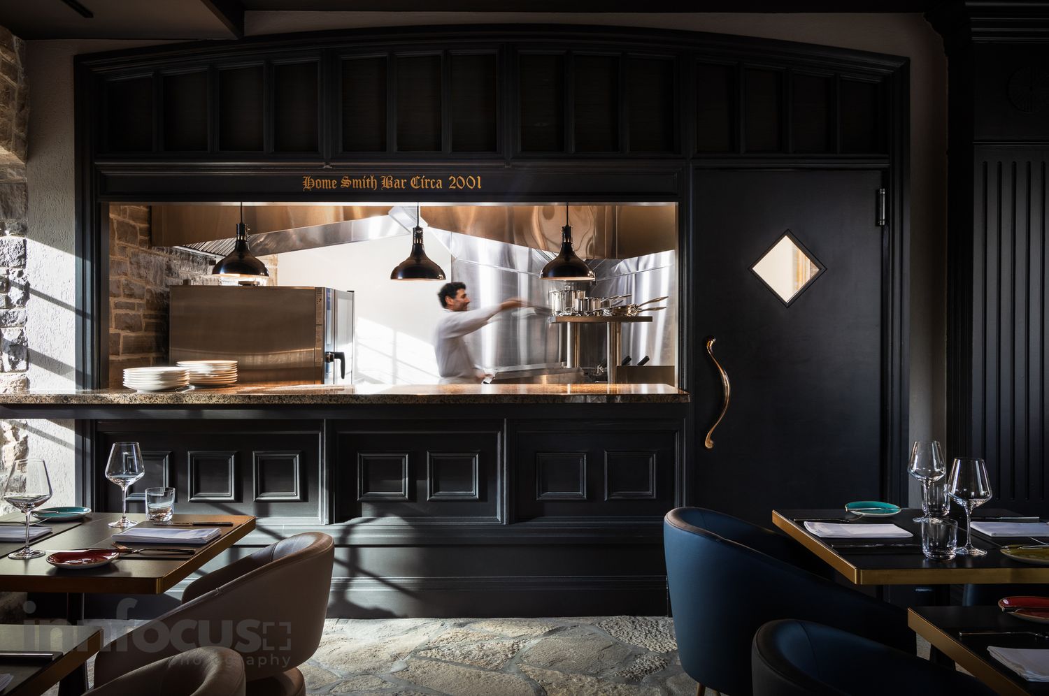 Upscale restaurant interior with dark walls, open kitchen view, pendant lights, and elegant table settings.