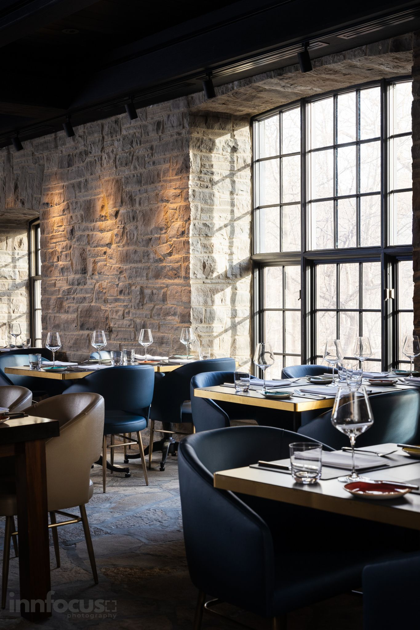Elegant restaurant interior with stone walls, large windows, blue leather chairs, and tables set with wine glasses.