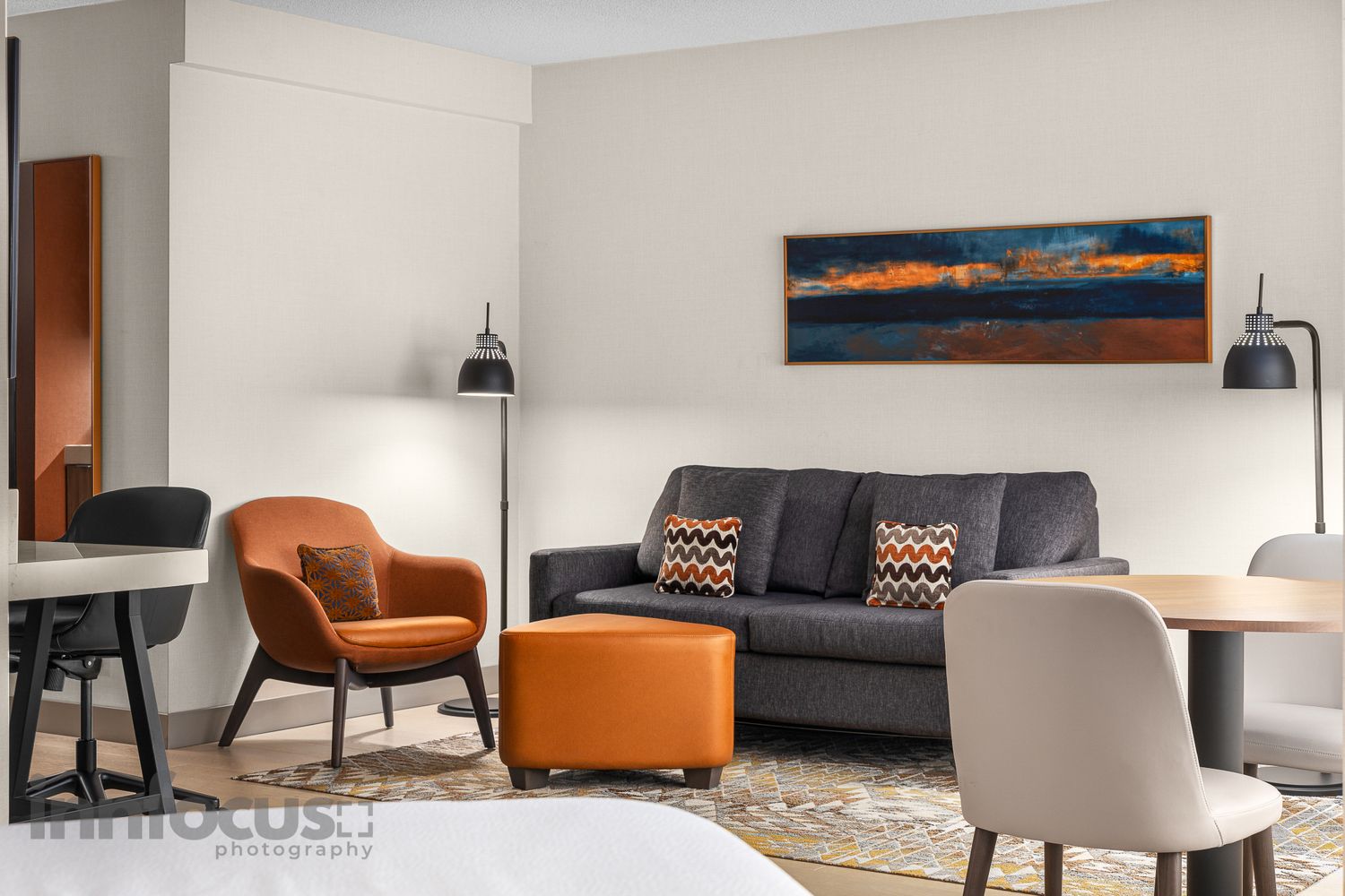 Modern hotel suite living area with gray sofa, orange armchair, ottoman, floor lamp, and landscape artwork on wall.