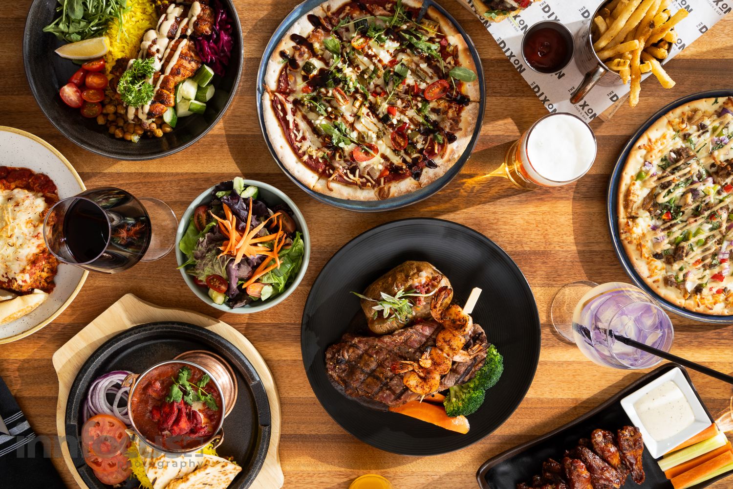 Overhead view of a restaurant spread featuring pizza, grilled steak, salads, pasta, and fries on a wooden table.