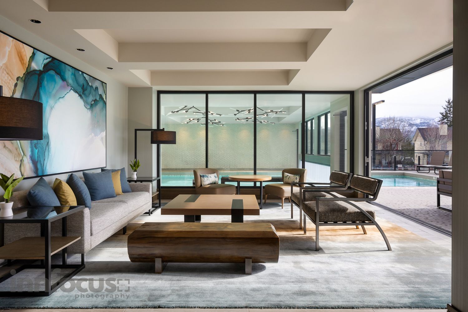 Modern luxury living room with large windows, wood coffee tables, colorful artwork, and a view of an outdoor pool.